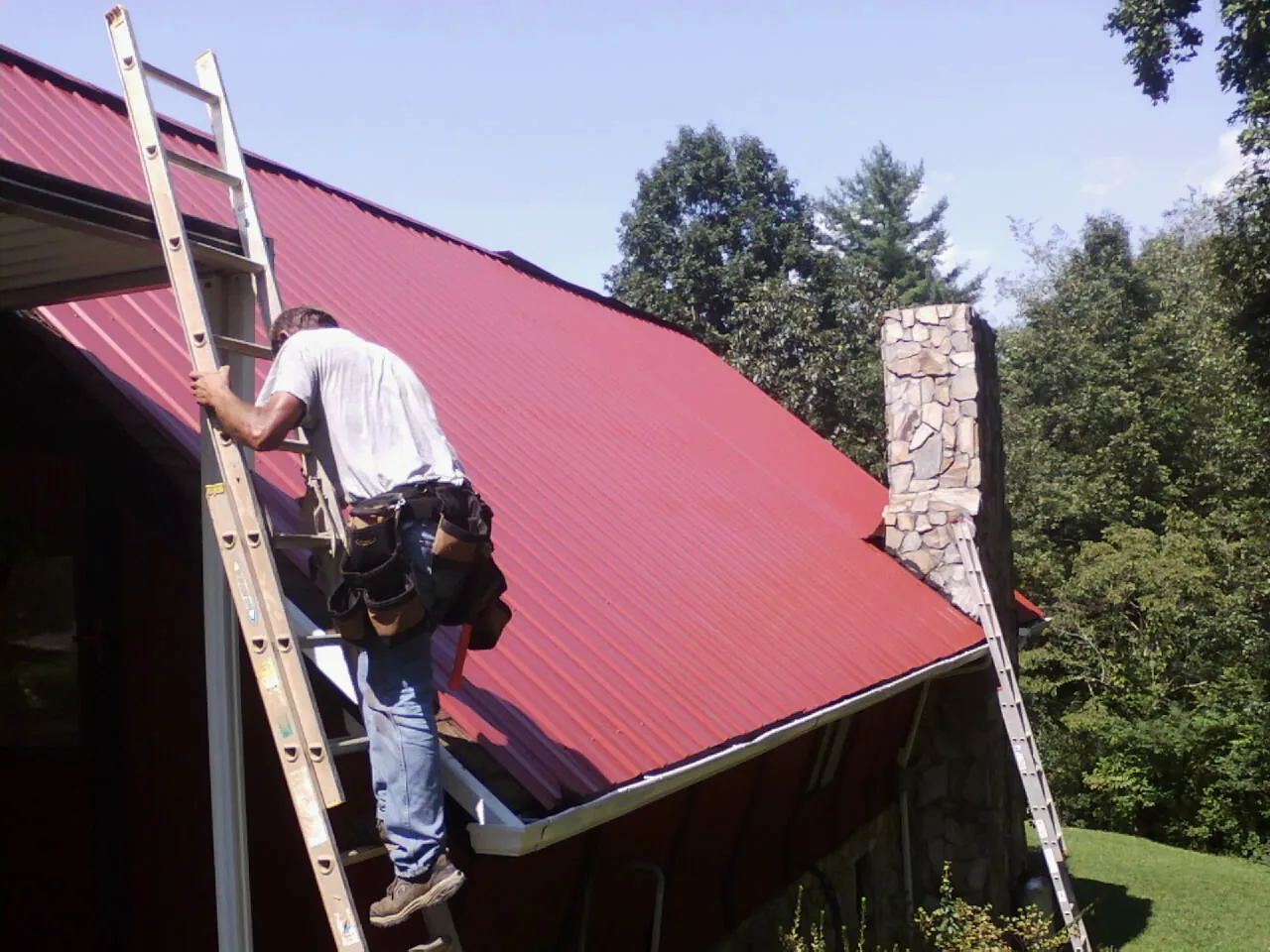 Roofer on a ladder beside a red metal roof and stone chimney — Soffit & Fascia Repair by Roofing Brothers in Fort Eustis